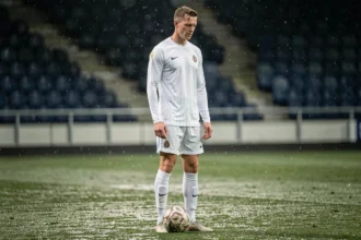 Footballeur hollandais introverti debout seul sur un terrain mouillé sous les tribunes vides, évoquant le style discret et génial de Dennis Bergkamp